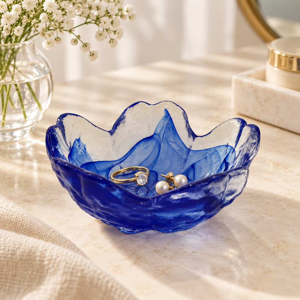 Blue glass flower-shaped trinket dish displayed on a cream vanity surface, holding a gold ring and pearl earrings, with soft flowers in the background.