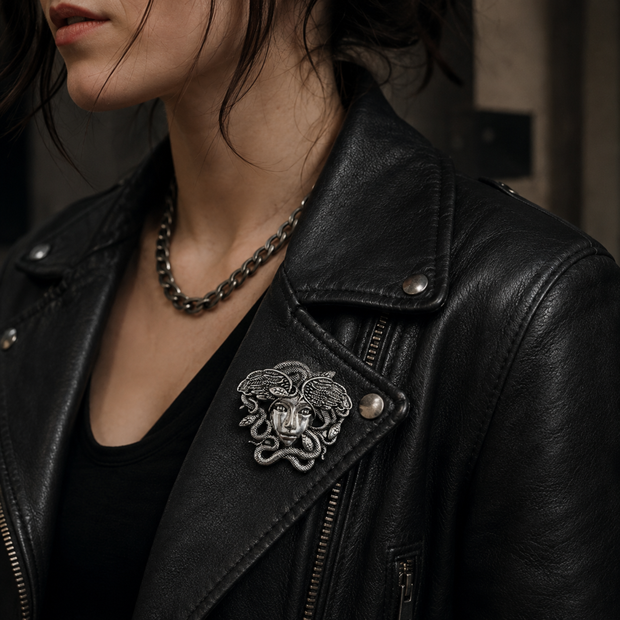 A close-up of a person wearing a black leather motorcycle jacket, featuring a prominent silver-tone Medusa head brooch pinned to the left chest, a dark chain necklace, and a black top underneath.