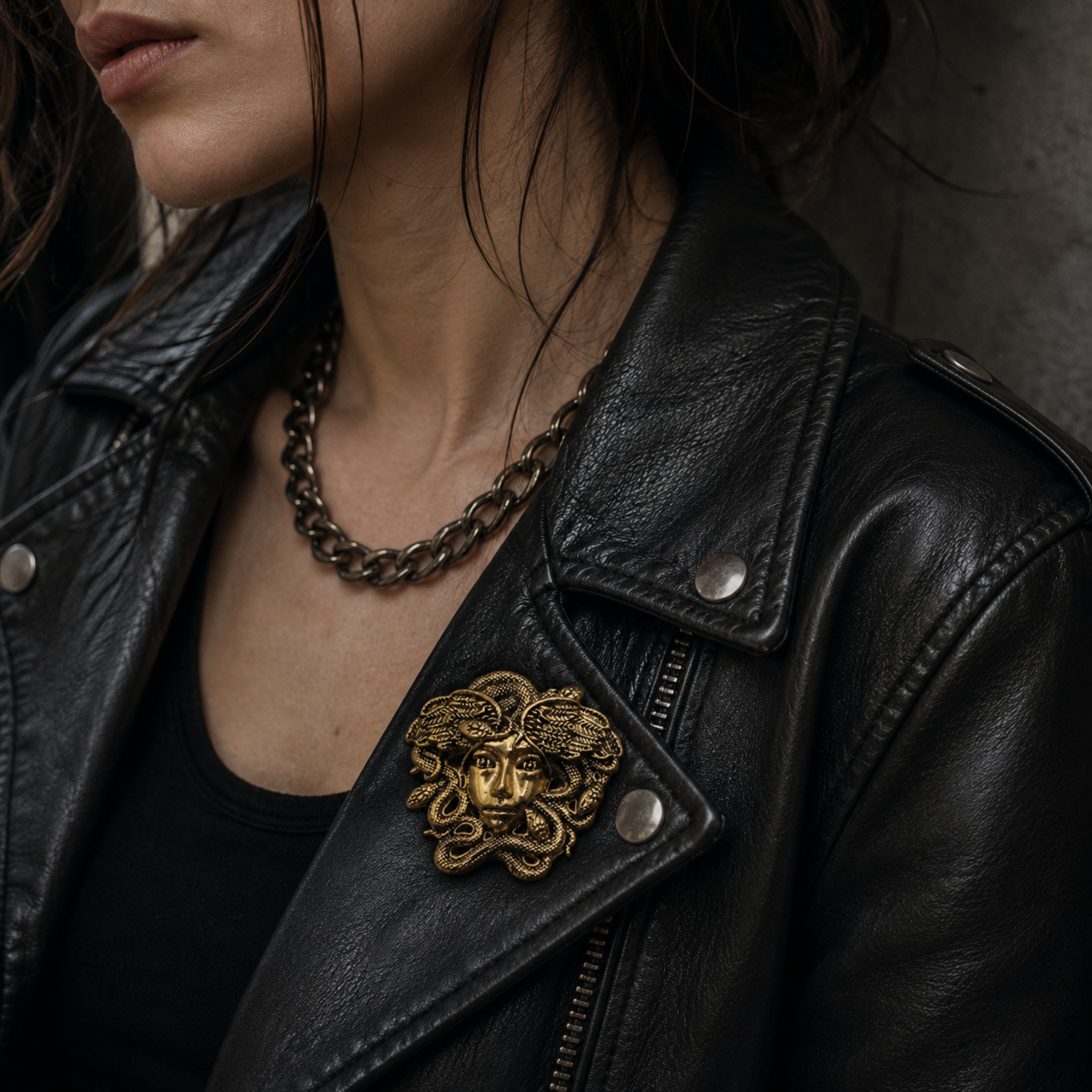 Close-up of a person wearing a black leather moto jacket with a large gold-tone Medusa head brooch pinned near the chest, along with a chunky chain necklace.