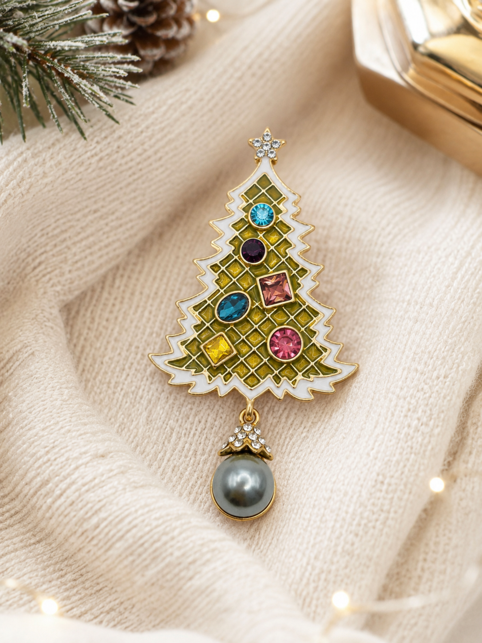 A gold-toned Christmas tree pendant brooch with colorful rhinestones and a dangling gray pearl, styled on a cream knit sweater with holiday greenery and warm lights.