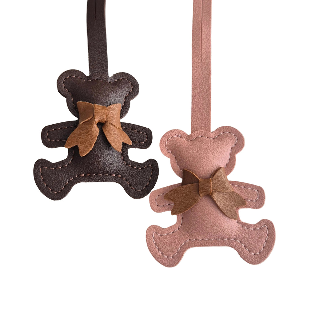 Two small teddy bear-shaped leather accessories hanging against a plain white background, one dark brown with a tan bow and one light pink with a tan bow.