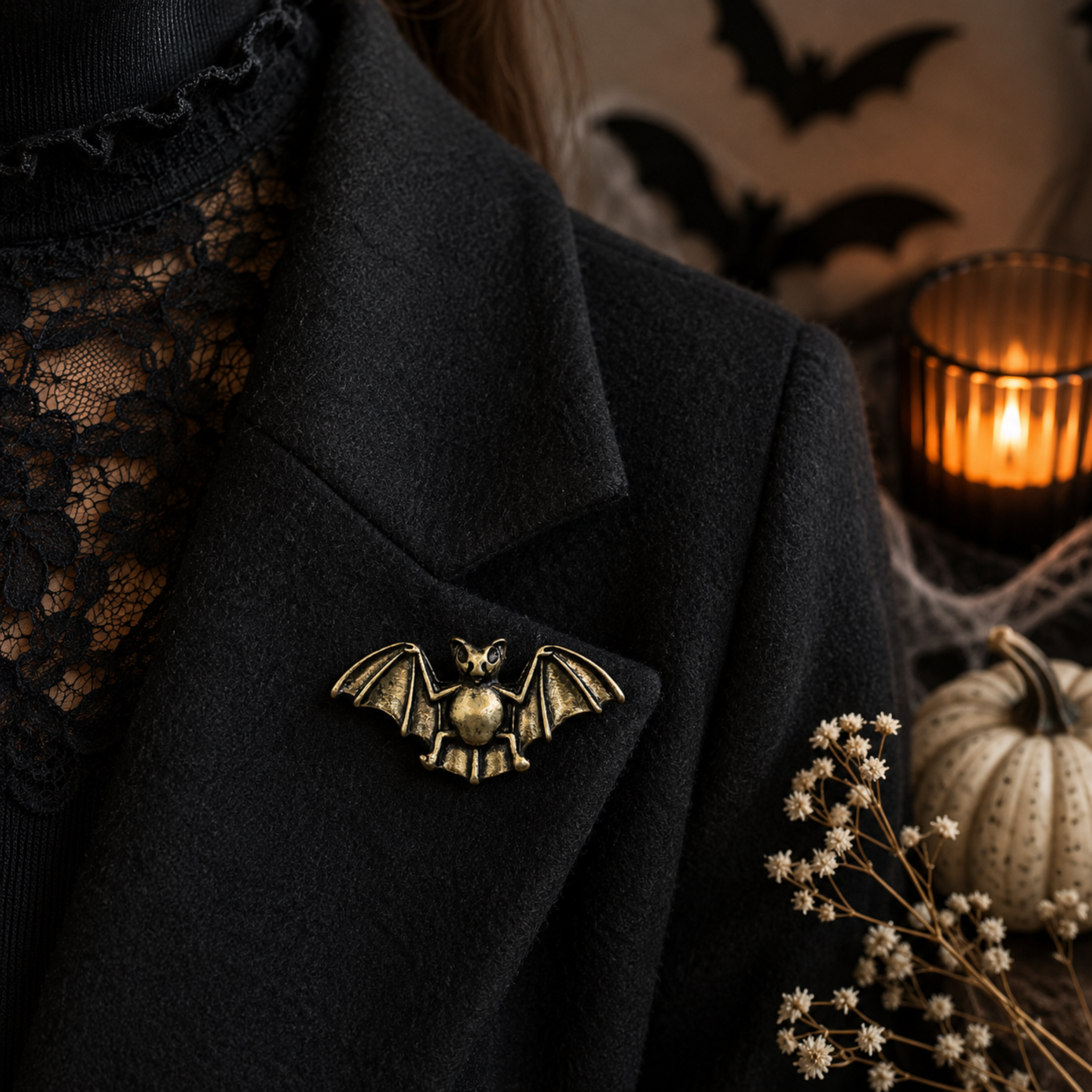 A close-up of a person wearing a black coat over a black lace top, with a small bat-shaped brooch pinned to the lapel. The background is dark and moody, with a lit candle, dried flowers, a small pumpkin, and bat silhouettes.