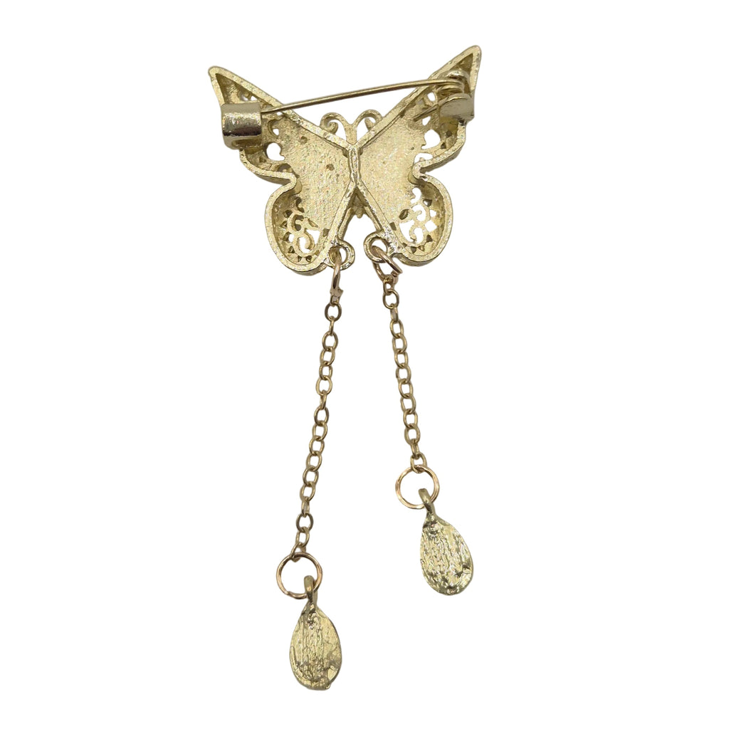 A gold butterfly-shaped brooch with intricate filigree detailing on the wings, featuring two dangling chain ends with small teardrop charms, photographed on a plain light gray background.