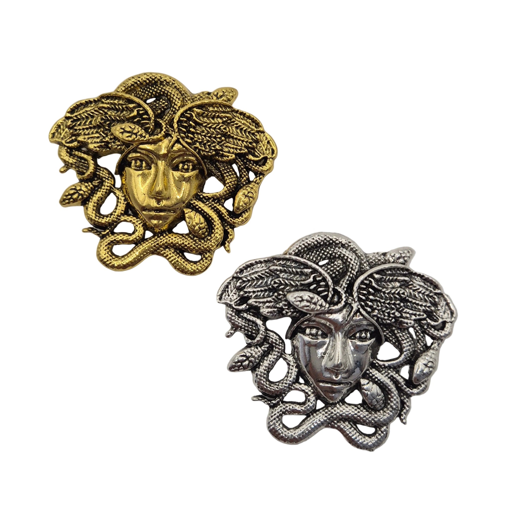 Two Medusa head brooches or charms with snakes, one in gold-tone metal and the other in silver-tone metal, displayed on a plain white background.
