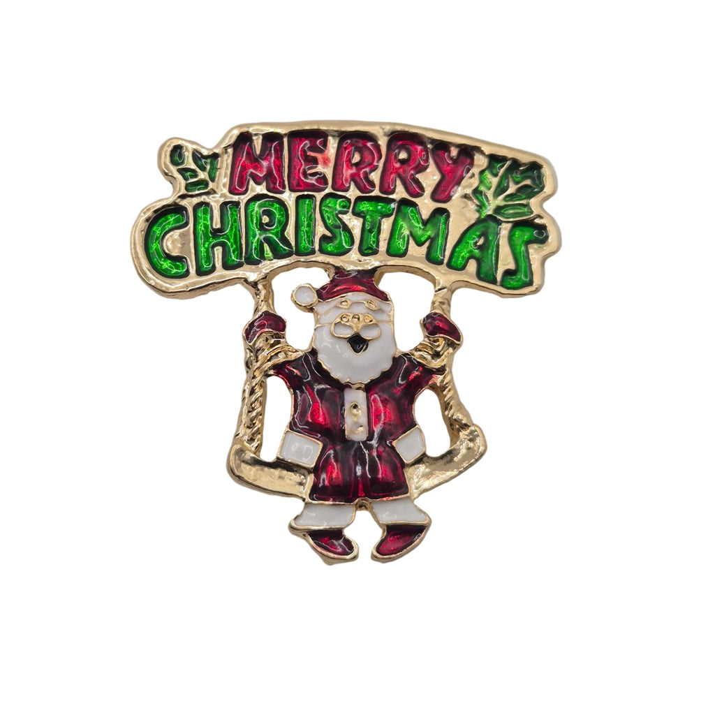 A Christmas-themed enamel pin featuring a gold-tone Santa Claus with a banner that reads 'MERRY CHRISTMAS' in green and red lettering, set against a plain white background. 