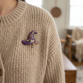 A purple witch hat brooch with gold-tone metal trim, decorated with small gold stars, rhinestone-embellished bands, and a dangling gold star charm, shown on a beige chunky knit cardigan.