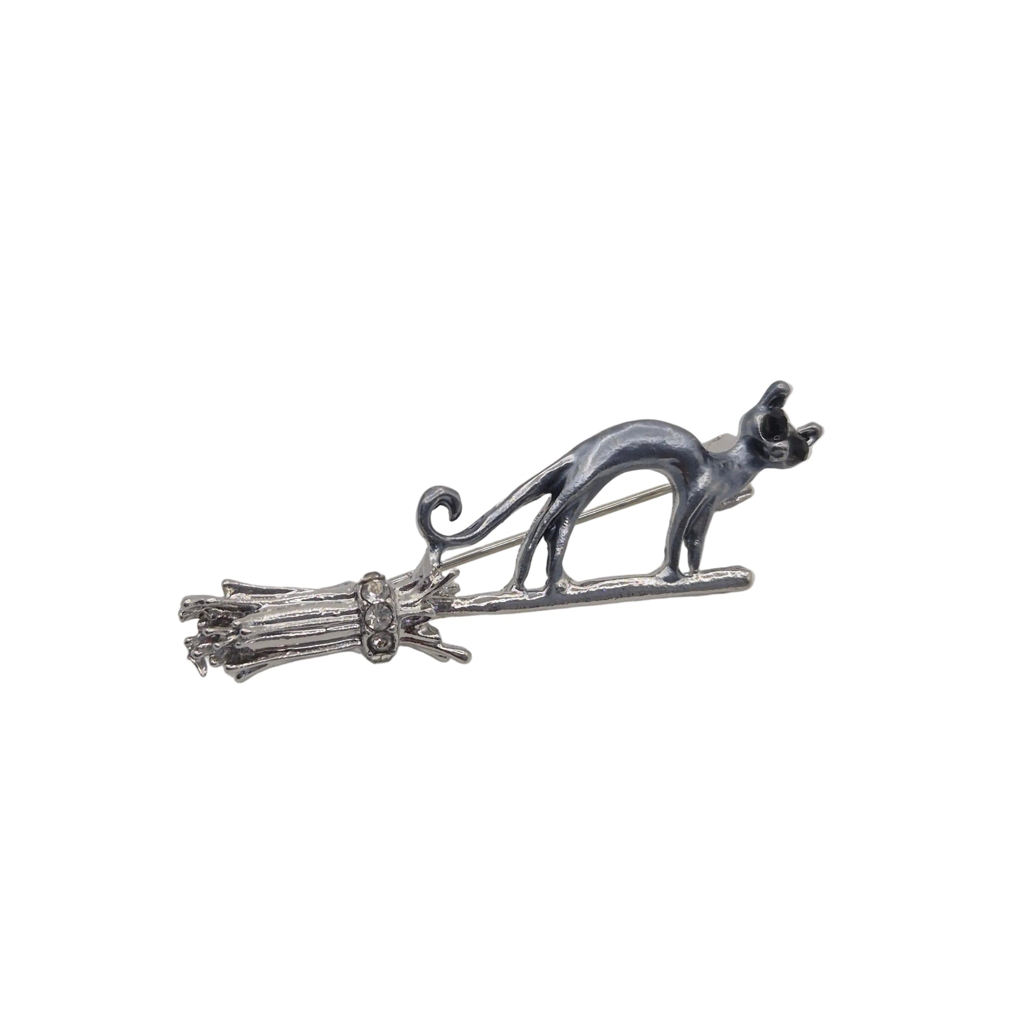 A silver-toned metal brooch featuring a cat standing on a broomstick, photographed against a plain white background.