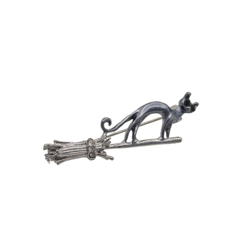 A silver-toned metal brooch featuring a cat standing on a broomstick, photographed against a plain white background.
