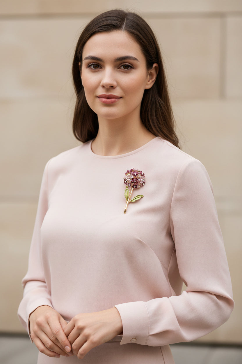 A gold-tone floral brooch featuring a round cluster of small flowers in pink, burgundy, and white enamel with clear rhinestones, and green enamel leaves on a stem. Shown on pink blouse. 