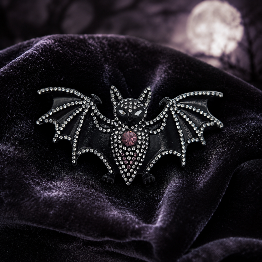 A gothic bat brooch embellished with clear rhinestones and a central pink gemstone, displayed on a dark purple velvet fabric with a blurred moonlit background.
