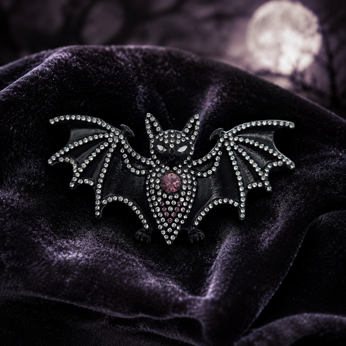 A gothic bat brooch embellished with clear rhinestones and a central pink gemstone, displayed on a dark purple velvet fabric with a blurred moonlit background.