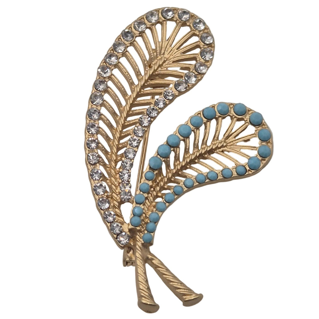 A gold-toned brooch shaped like two overlapping stylized feathers, decorated with clear rhinestones on one leaf and small turquoise blue stones on the other, shown on a plain white background.