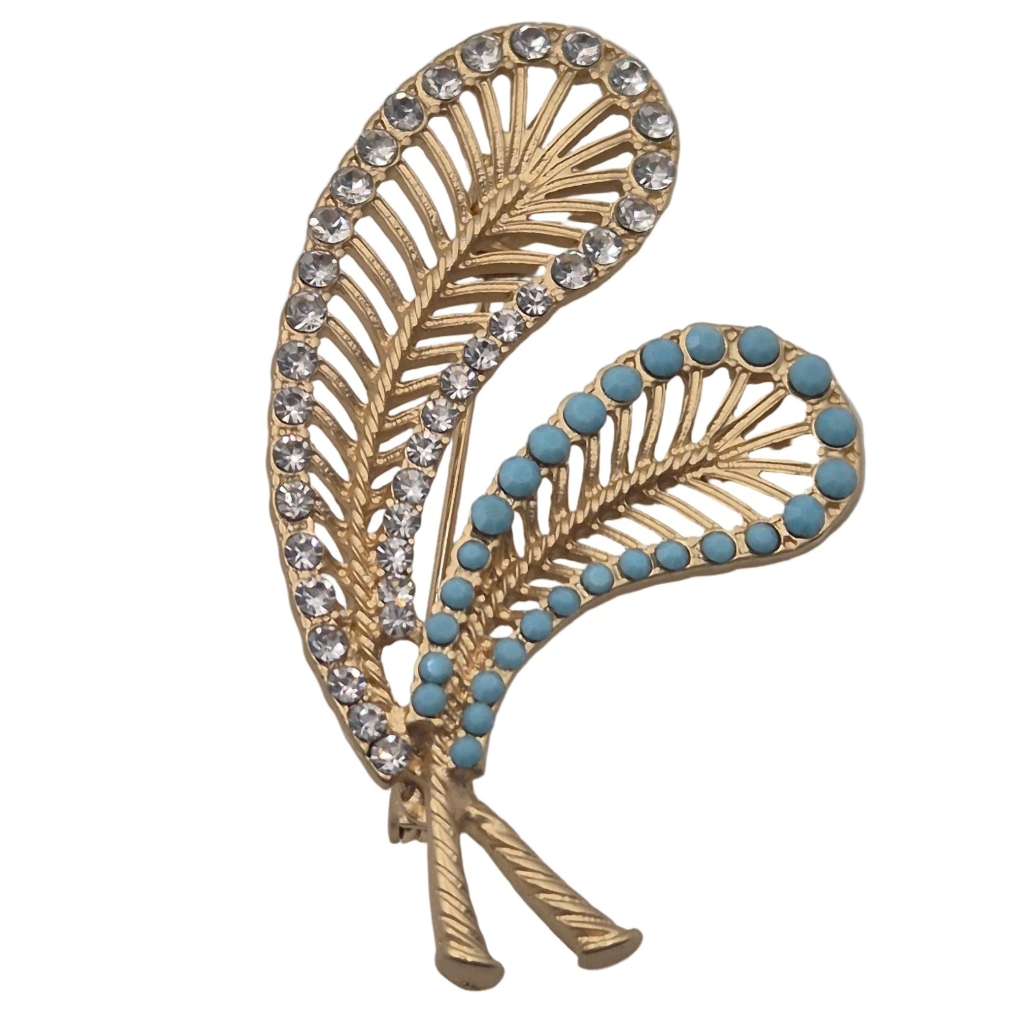 A gold-toned brooch shaped like two overlapping stylized feathers, decorated with clear rhinestones on one leaf and small turquoise blue stones on the other, shown on a plain white background.