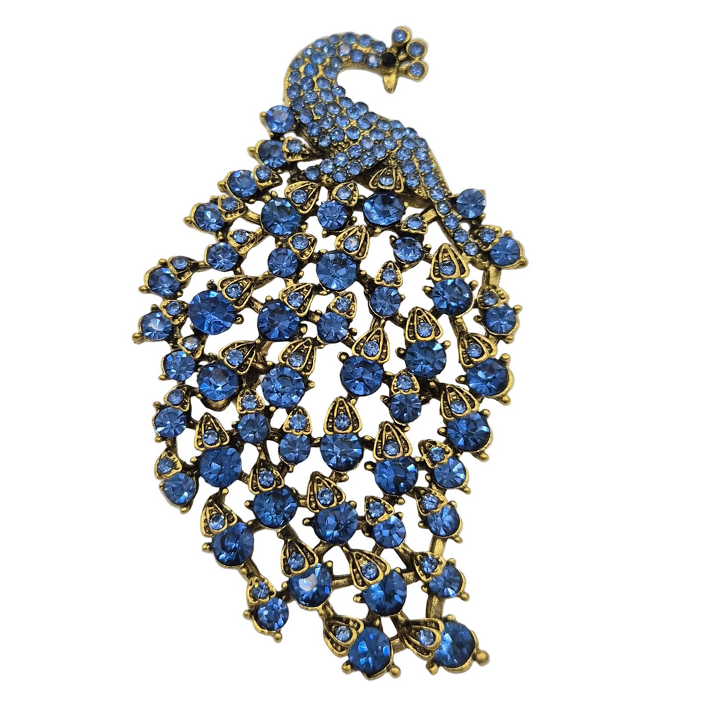 A peacock brooch featuring a gold-tone metal body adorned with numerous round and teardrop-shaped blue rhinestones, set against a plain white background.