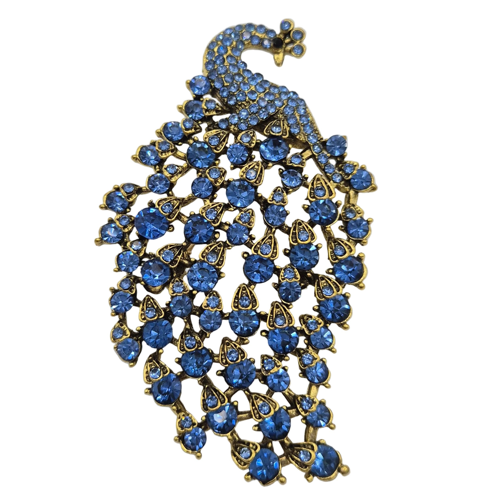 A peacock brooch featuring a gold-tone metal body adorned with numerous round and teardrop-shaped blue rhinestones, set against a plain white background.