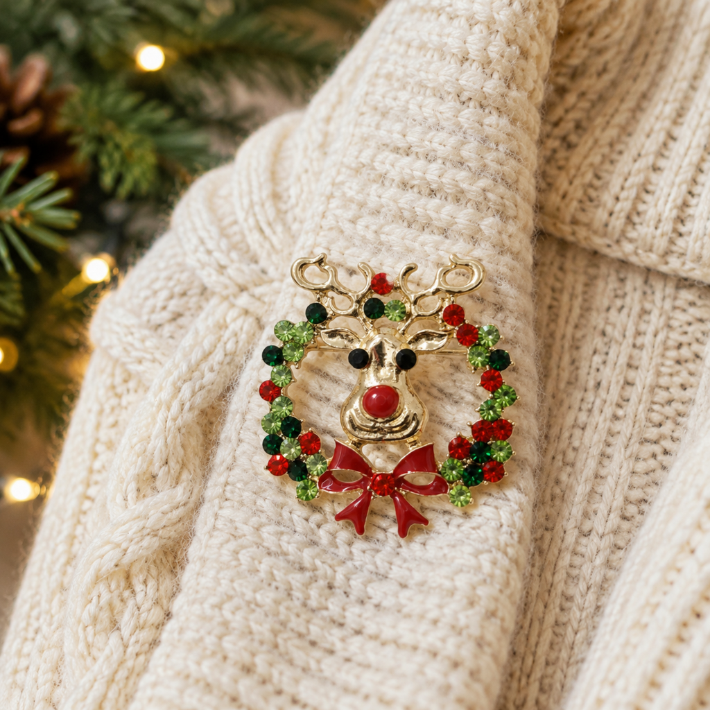 A gold-toned Christmas reindeer brooch with a red nose and antlers, surrounded by a green and red beaded wreath and a red bow, pinned onto a cream cable-knit sweater.