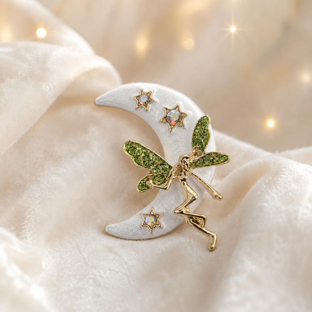 A fairy and crescent moon brooch with sparkling green wings and gold-toned details, displayed on a soft cream fabric background with warm bokeh light effects.