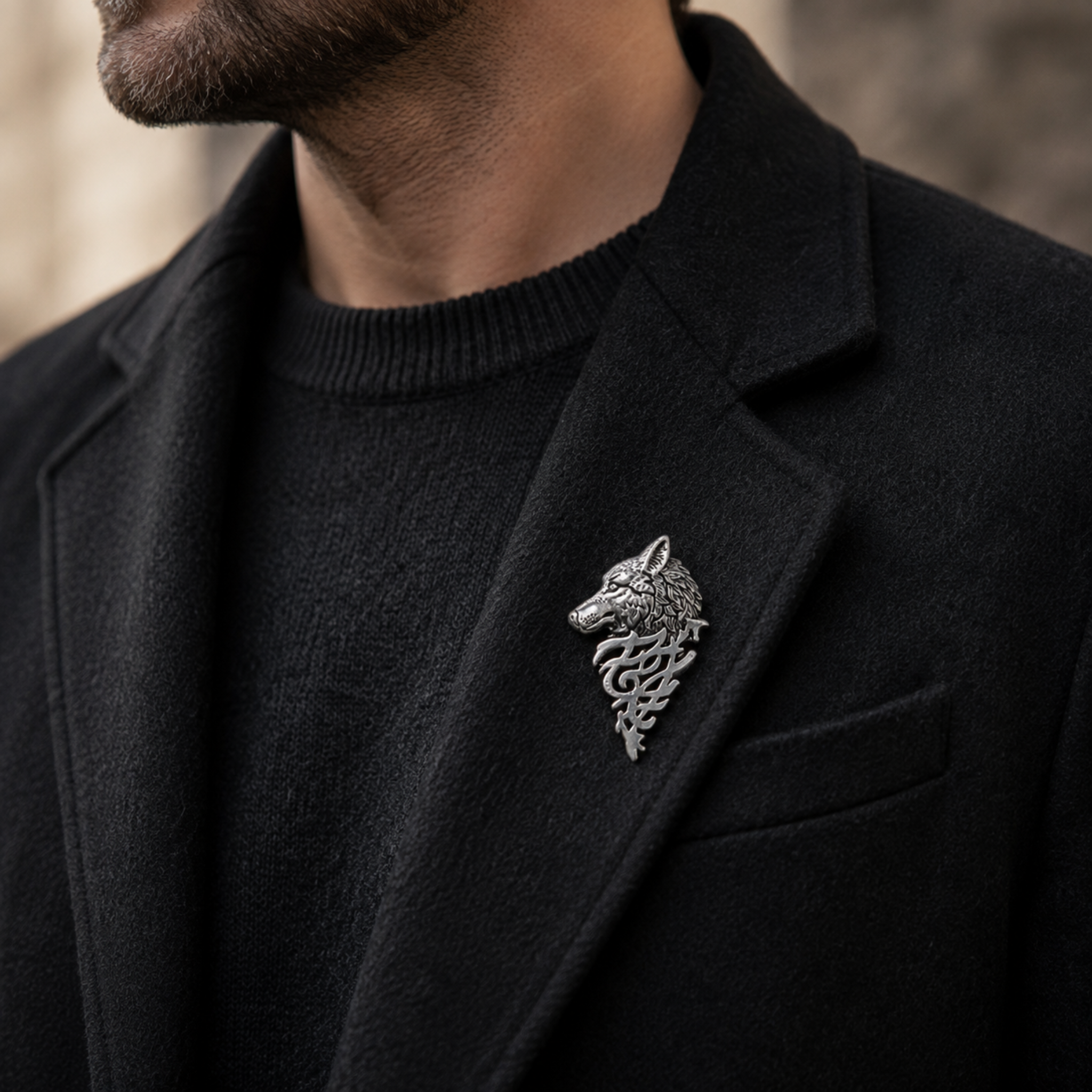 A silver-toned metal wolf head brooch with intricate scrollwork, pinned to the lapel of a black wool blazer worn over a black knit sweater.