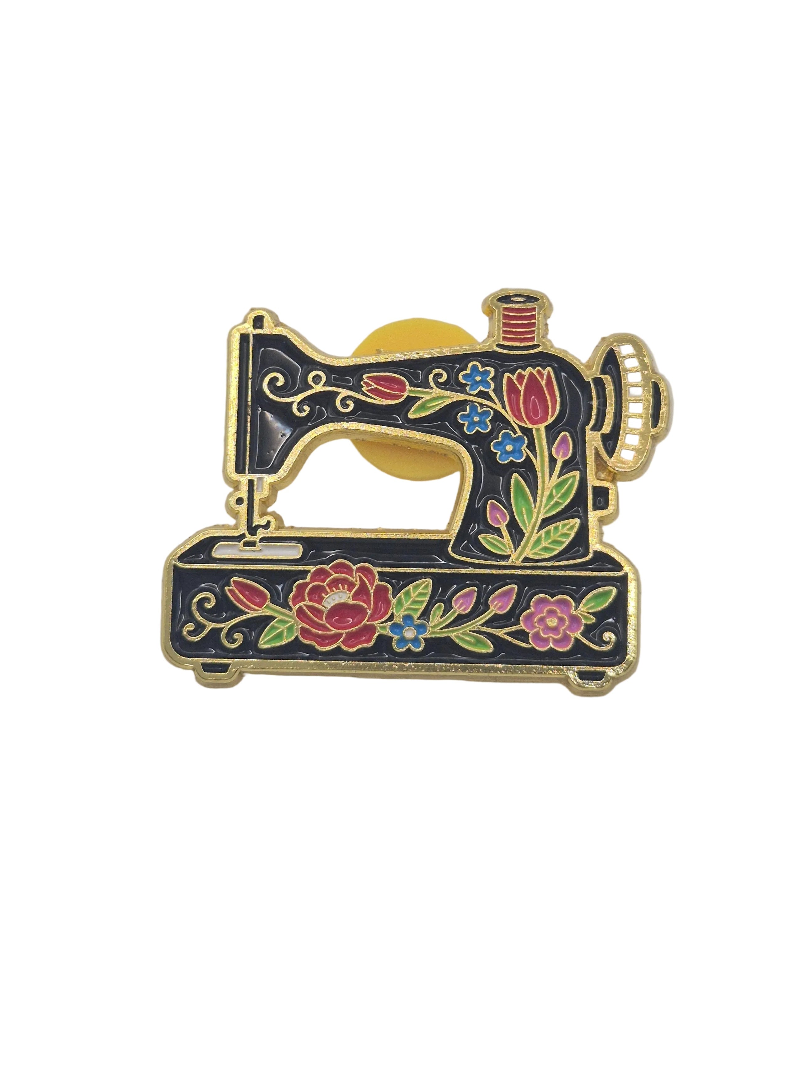A decorative enamel pin shaped like a sewing machine, featuring a black body with gold-tone metal outlines and colorful floral details in pink, red, blue, and green, set against a plain white background.