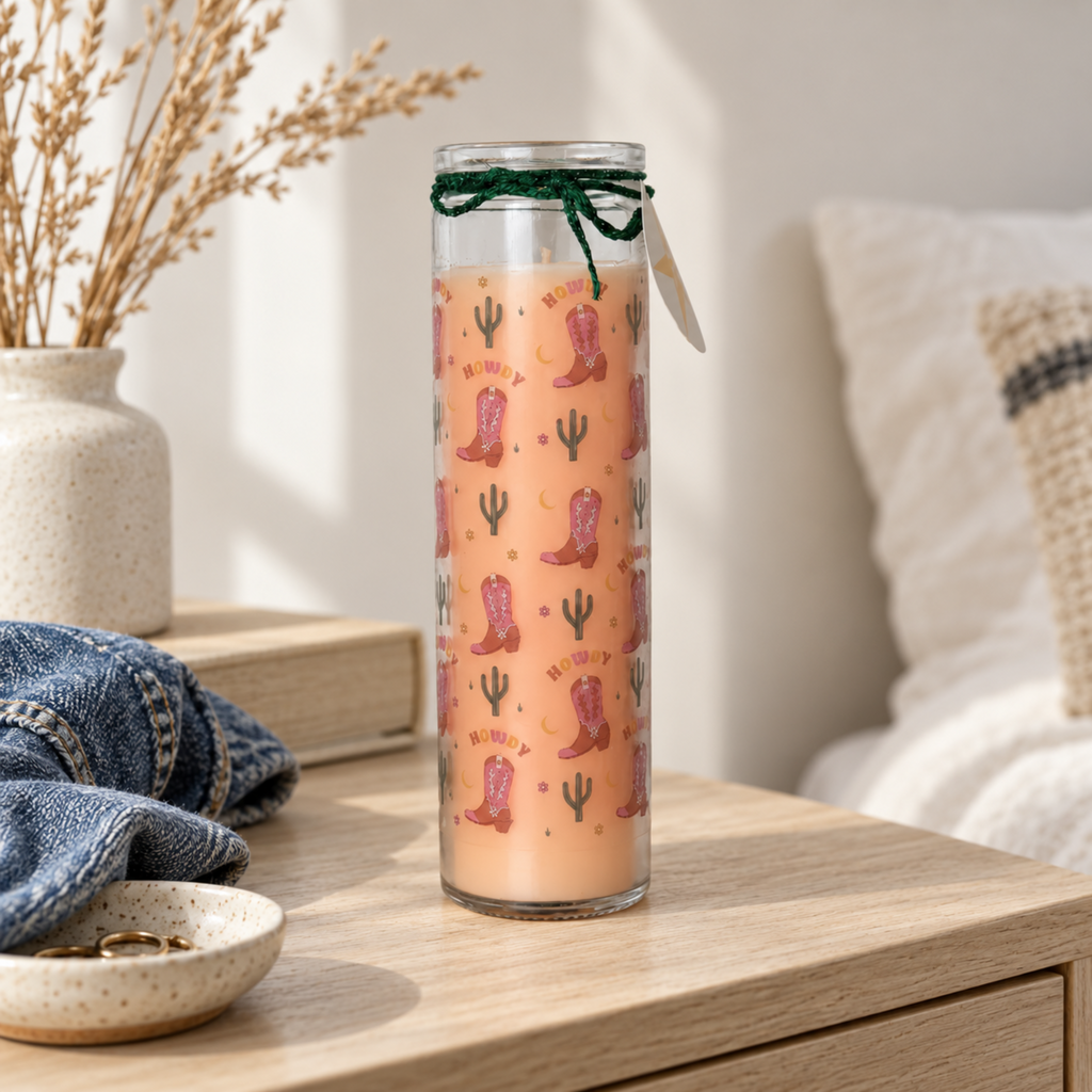 A tall glass jar candle with peach-coloured wax, decorated with a pink and green western boot and cactus pattern, displayed on a wooden side table with flowers in the corner.