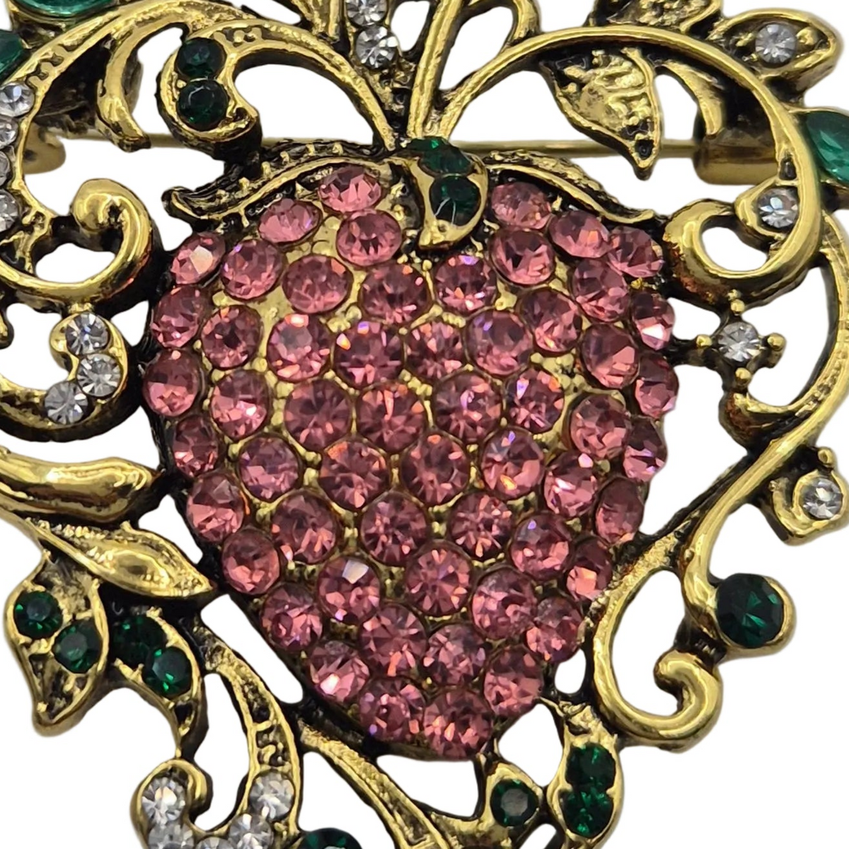 A close-up of an ornate brooch featuring a heart-shaped center paved with pink rhinestones, surrounded by gold-tone scrollwork with small green and clear rhinestones on a white background.
