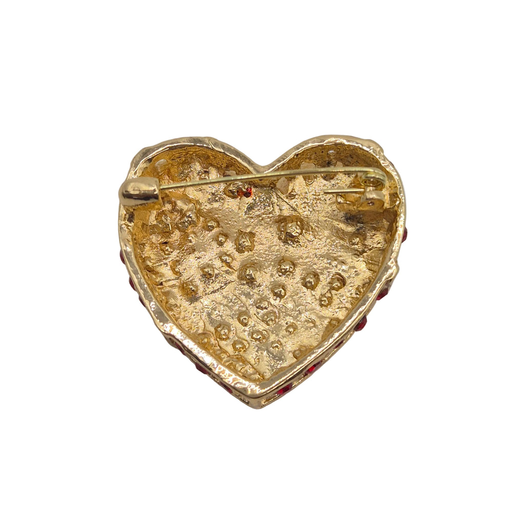 A gold heart-shaped brooch with a textured, dotted surface and a safety pin clasp, accented with small red gemstones along the edge, shown on a neutral white background.