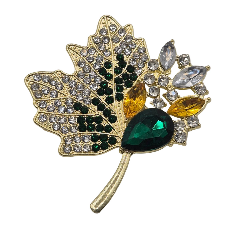 A gold-toned maple leaf brooch embellished with clear rhinestones, dark green stones, and yellow gemstones on a white background.