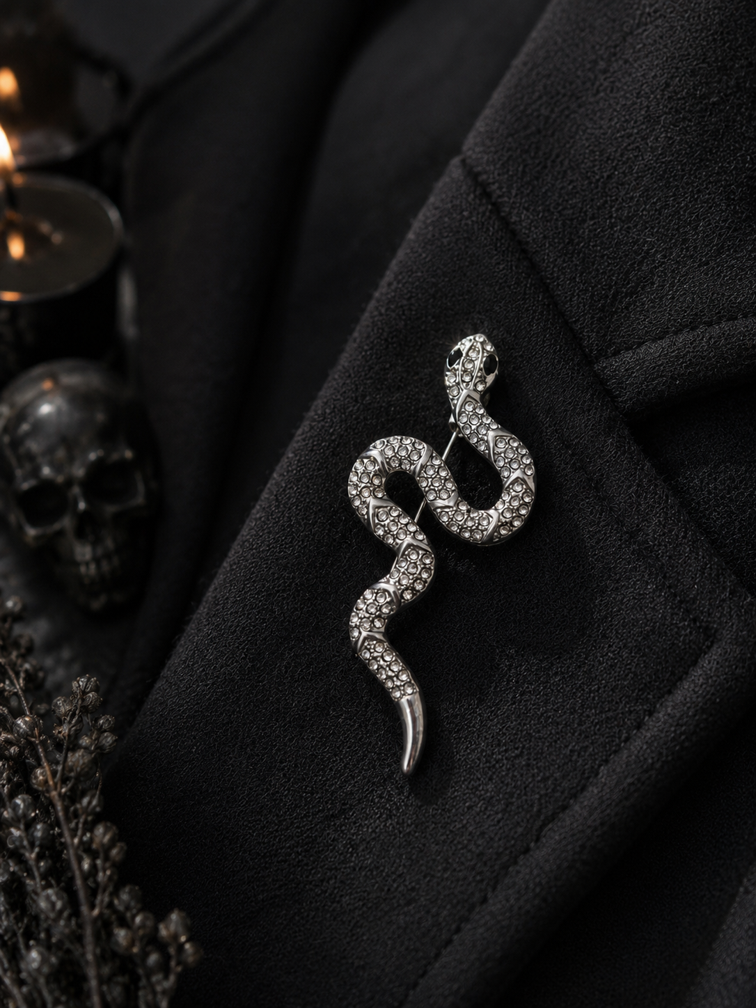 A silver snake-shaped brooch embellished with clear rhinestones, pinned to a black coat. The background features dark gothic props including a skull and dried flowers.