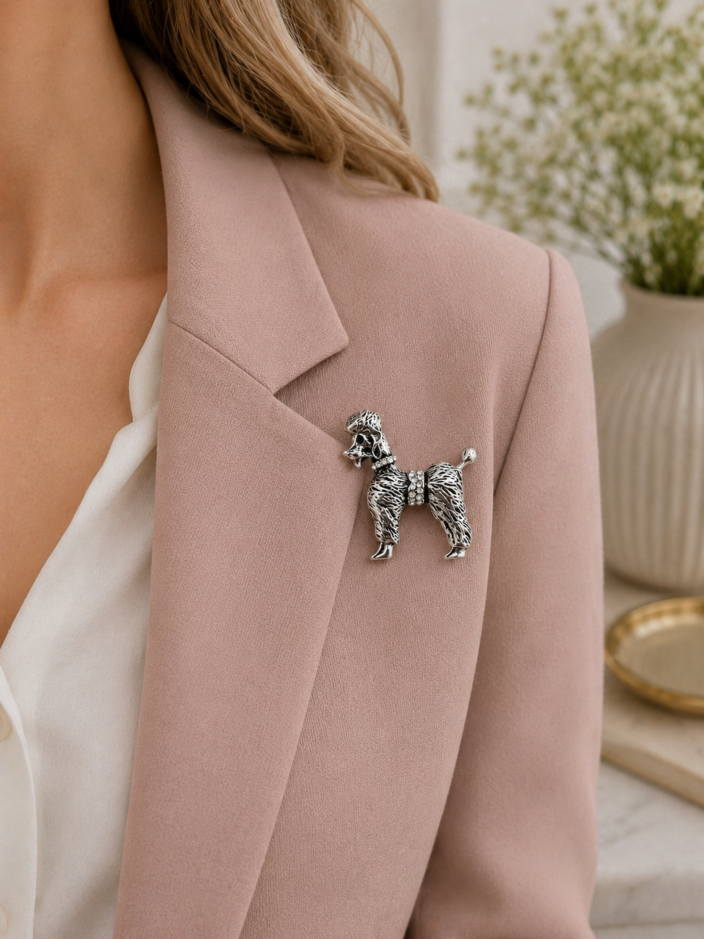 A silver poodle-shaped brooch pinned to the lapel of a dusty pink blazer worn by a model.