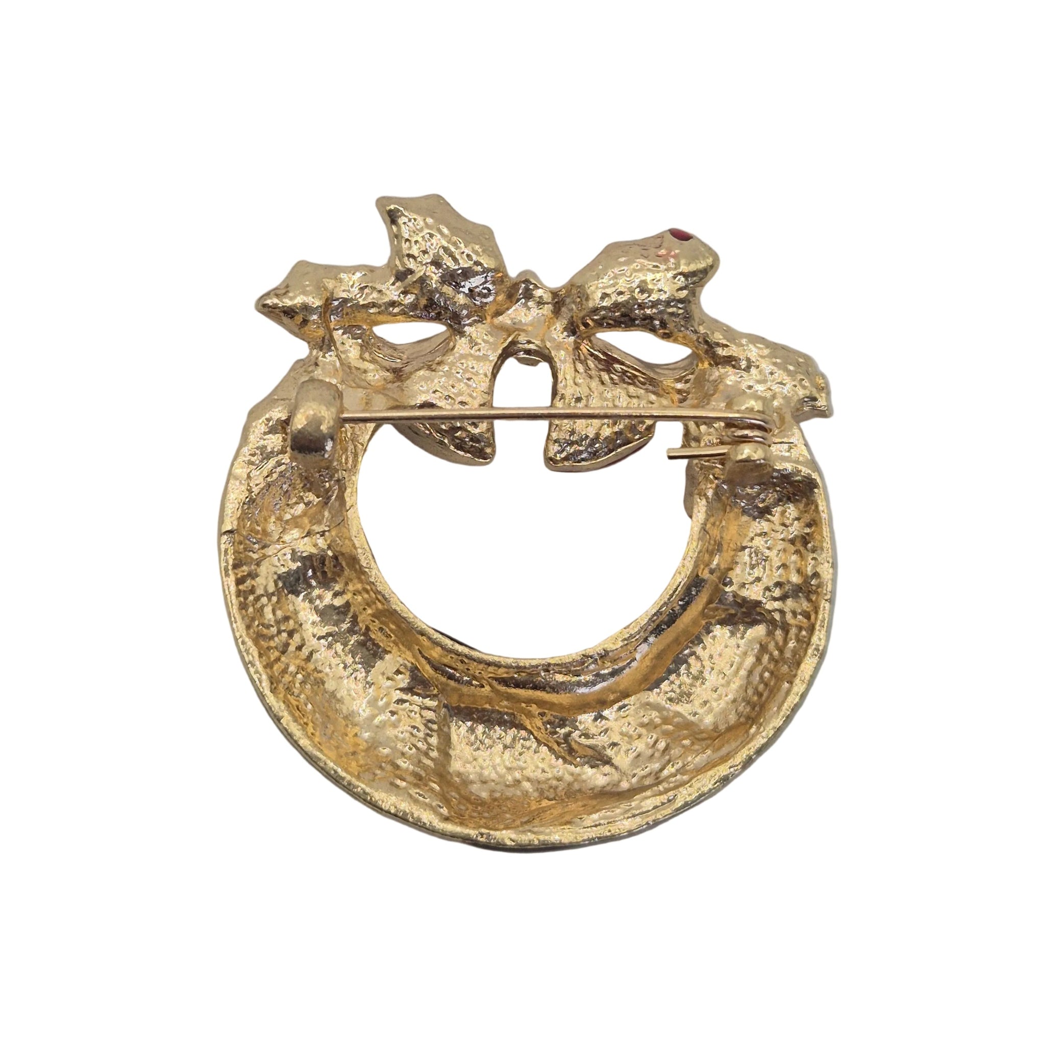 A textured gold-tone bow and circular ring brooch with a small red gemstone accent, photographed on a neutral light gray background.