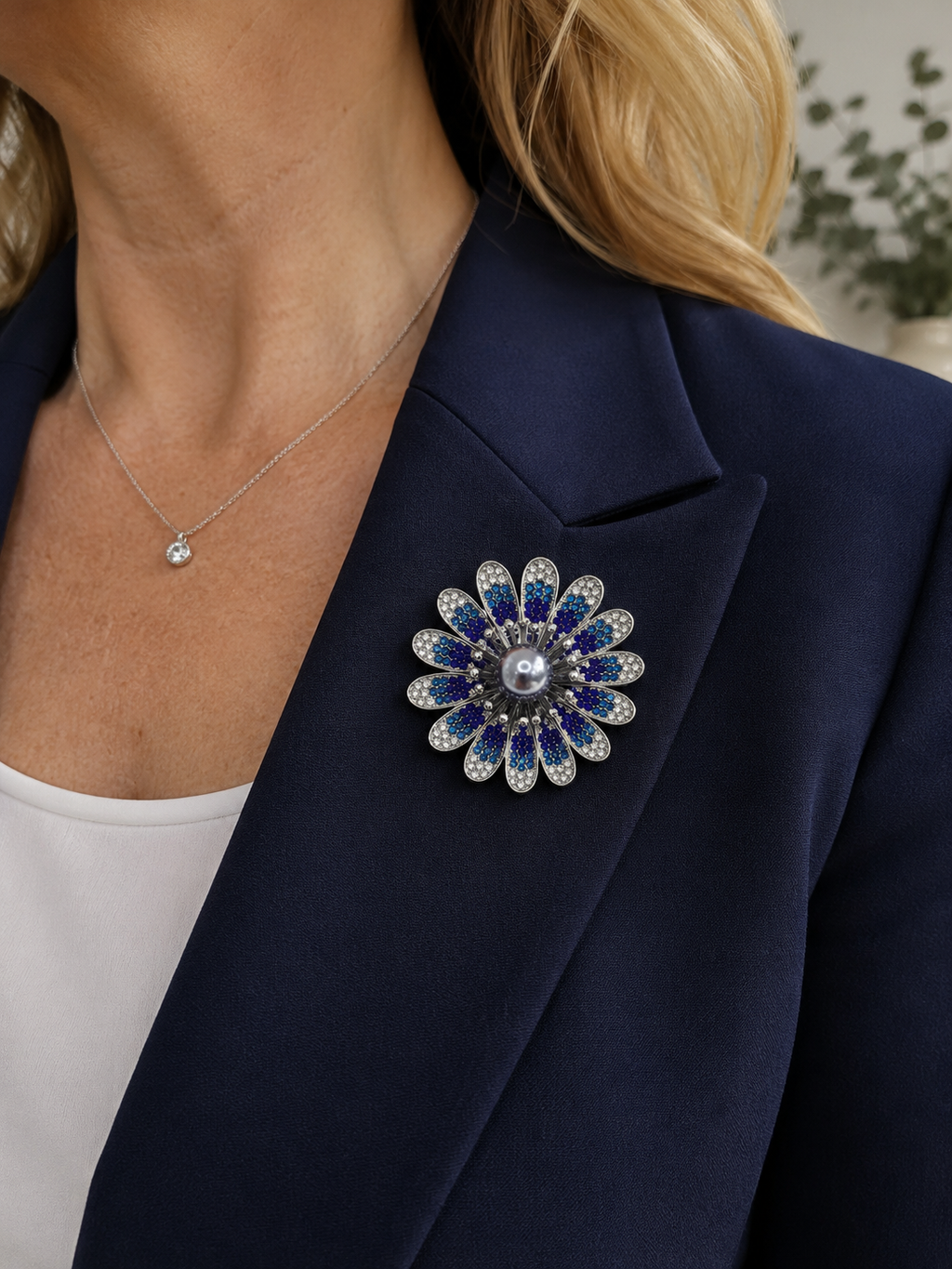 A close-up of a woman wearing a navy blue blazer, a delicate silver necklace, and a silver flower-shaped brooch embellished with deep blue and clear rhinestones.
