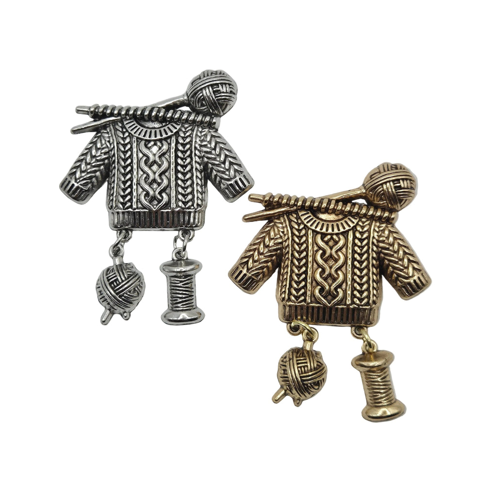 Two metal jumper brooch pins on a plain white background, one in silver-toned metal and the other in gold-toned metal, both detailed with knit patterns, yarn balls, and spools.