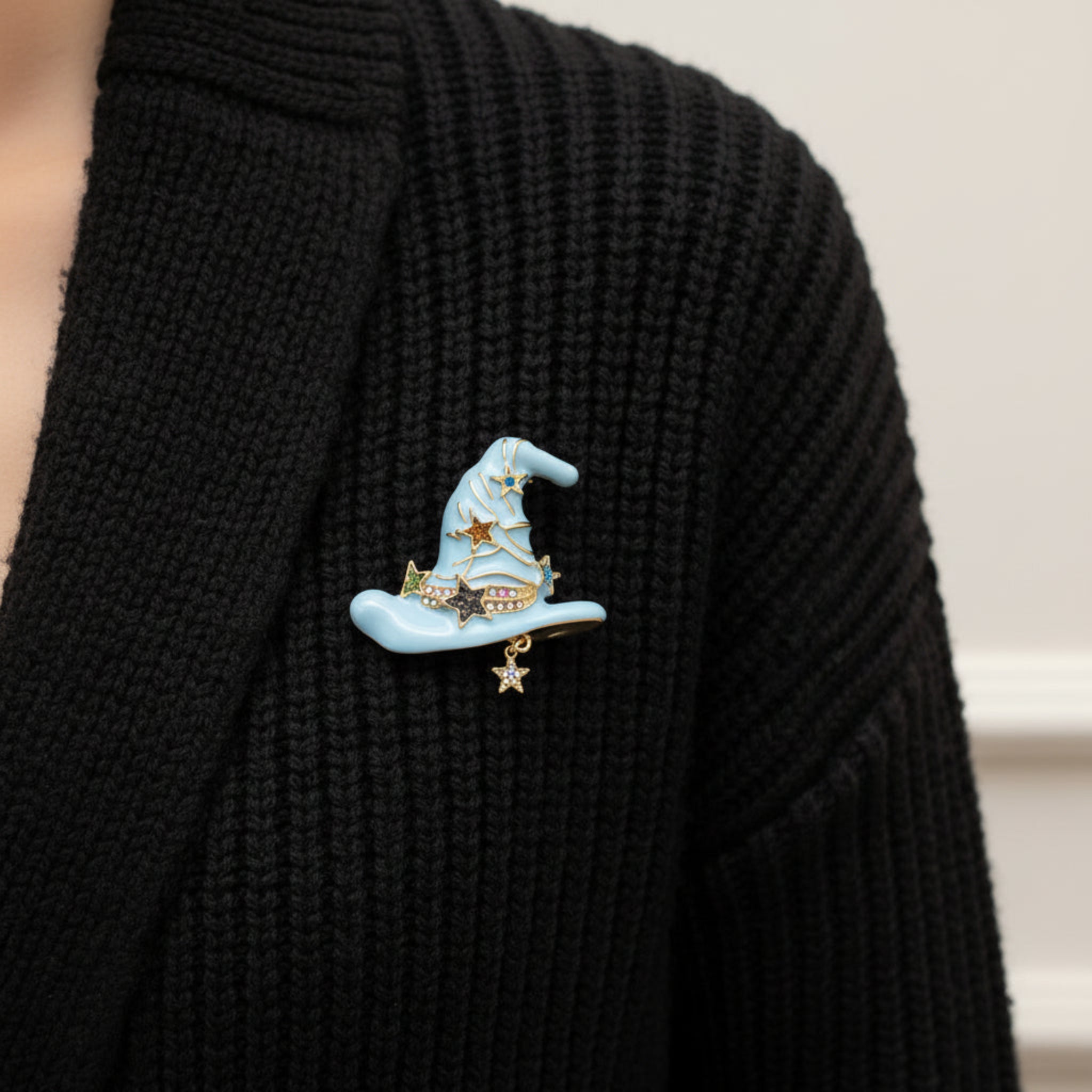 A light blue witch hat brooch with gold-tone metal trim, decorated with gold, brown, and blue star charms, and a dangling star charm at the bottom, shown on a black chunky knit cardigan.