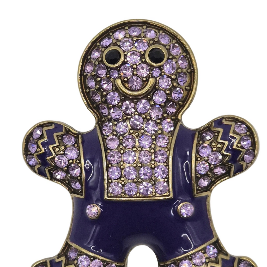 A gingerbread man-shaped brooch pin, featuring a dark purple enamel body, gold metal trim, and light purple rhinestones covering the head, arms, and torso.