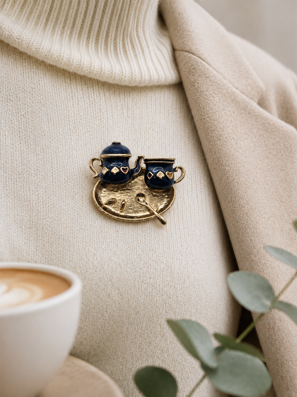 A close-up of a gold-toned enamel lapel pin shaped like a coffee pot and cup, pinned onto a beige jumper with coat over. Small green leaves appear in the lower right corner.