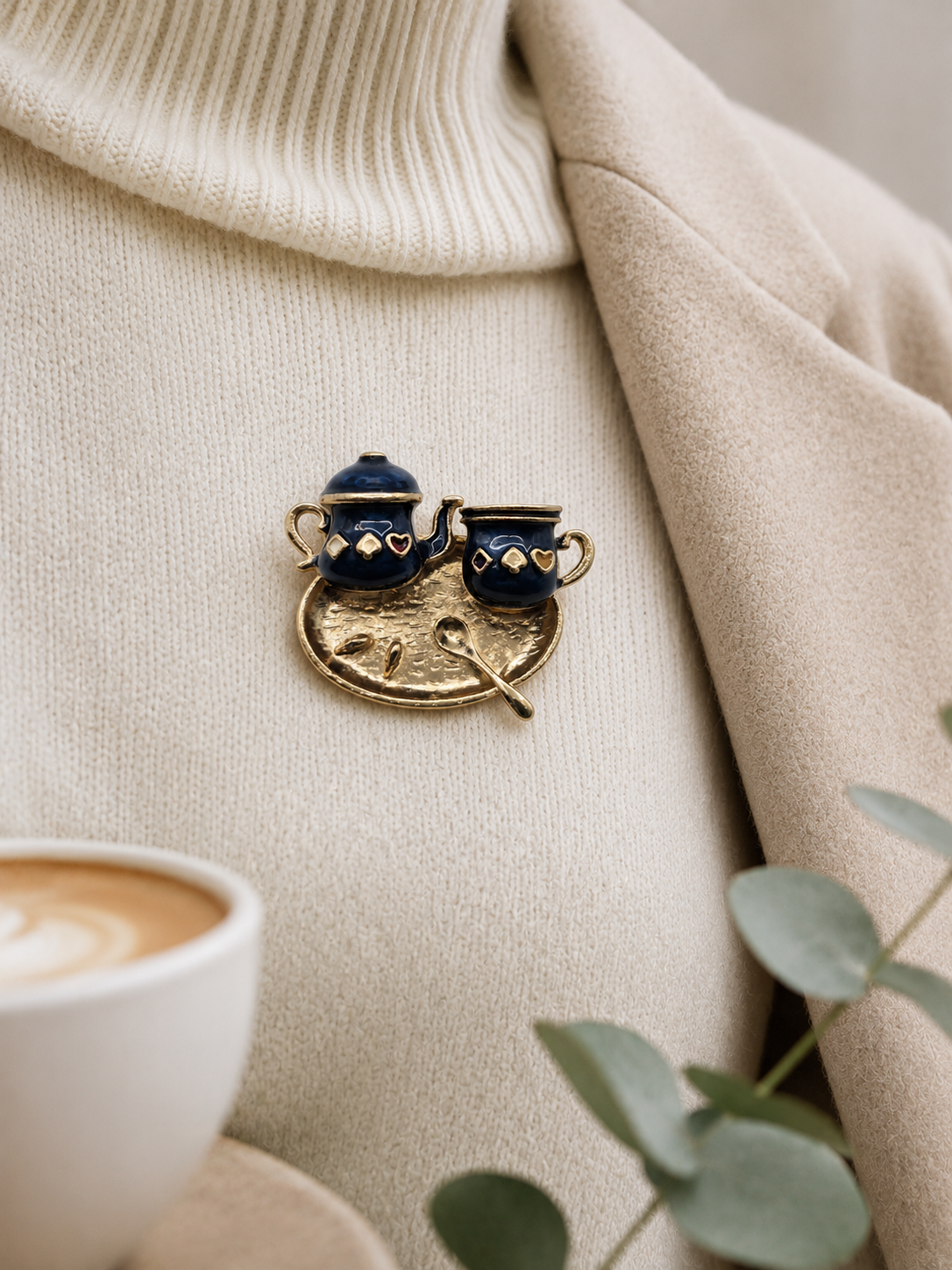 A close-up of a gold-toned enamel lapel pin shaped like a coffee pot and cup, pinned onto a beige jumper with coat over. Small green leaves appear in the lower right corner.