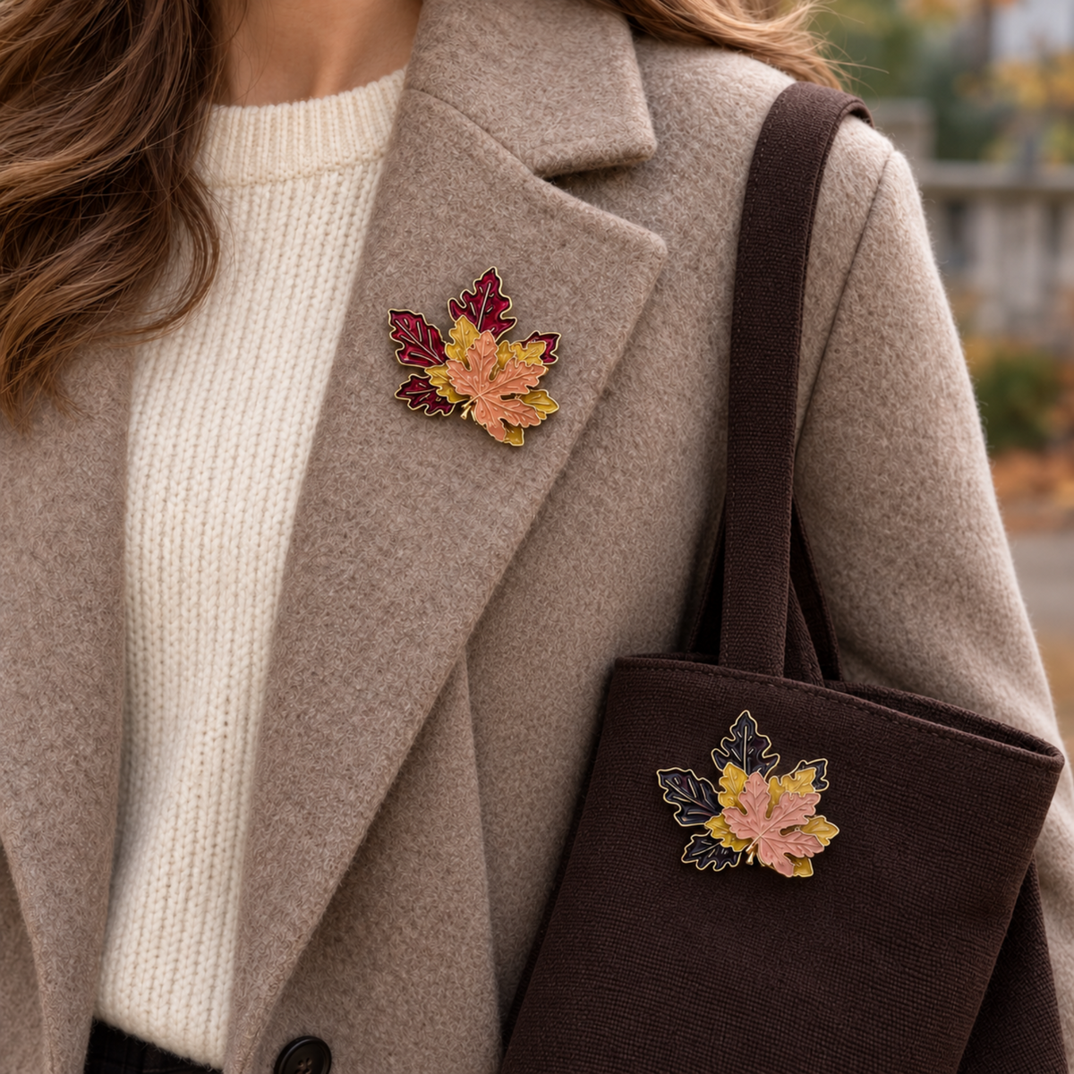 A person wearing a beige coat over a cream sweater carries a dark brown tote bag. Both the coat and bag feature a matching autumn leaf brooch in shades of pink, yellow, and red.