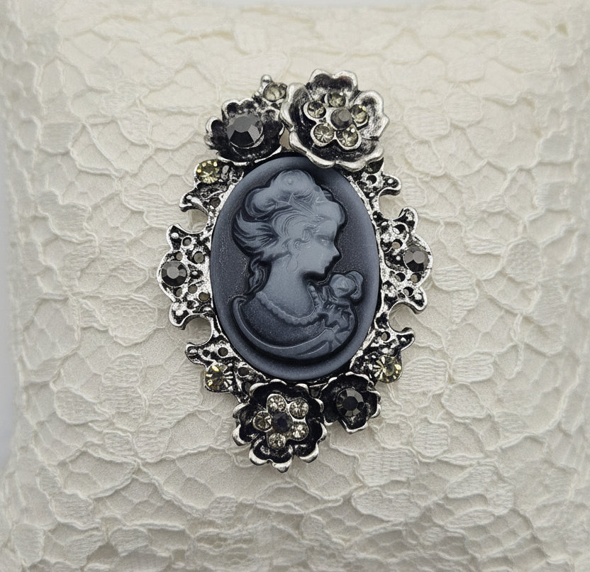 A vintage-style cameo brooch featuring a gray carved profile of a woman within an ornate silver-toned floral border, set against a textured white lace background.