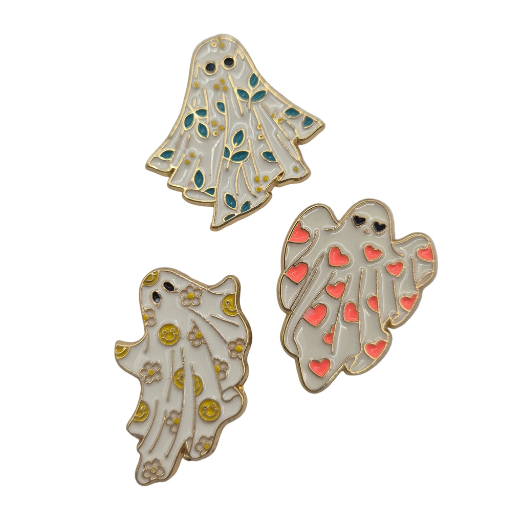 Three enamel ghost-shaped pins on a white background. One ghost has teal and green leaf patterns, one has yellow smileys and stars, and one has pink hearts.