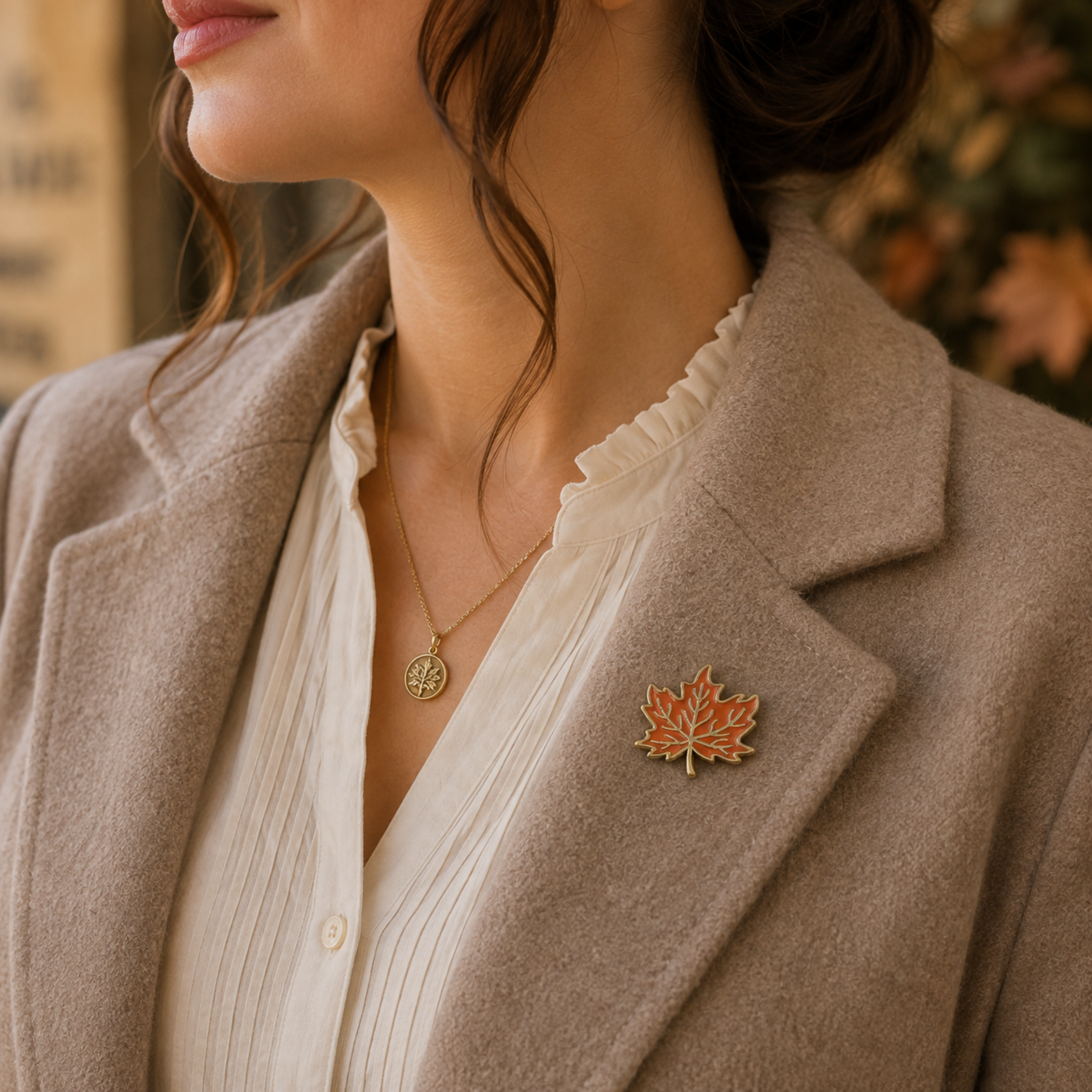 A close-up lifestyle shot of a woman wearing a gold maple leaf brooch with orange and gold enamel detailing on her beige blazer.