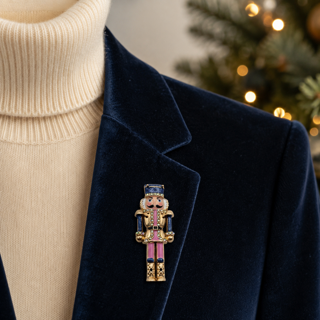 A close-up of a navy velvet blazer over a cream ribbed turtleneck sweater, featuring a decorative nutcracker pin on the lapel. The nutcracker pin has gold, pink, and blue details, with a softly blurred Christmas tree in the background.