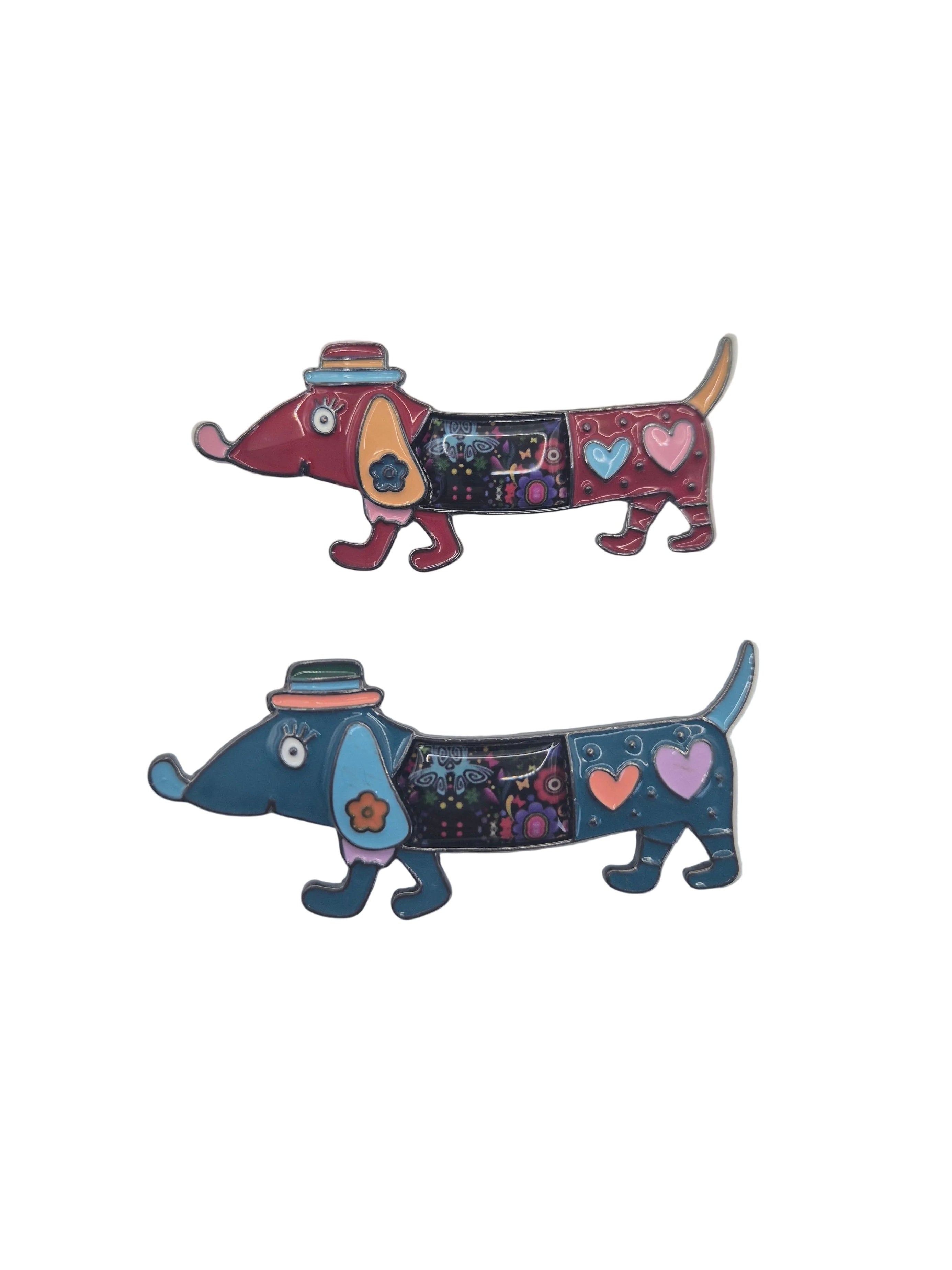 Two colorful brooches shaped like dachshund dogs, one in red and burgundy with a pink hat, and the other in blue and teal with a dark hat, both decorated with hearts and stars, on a plain white background.
