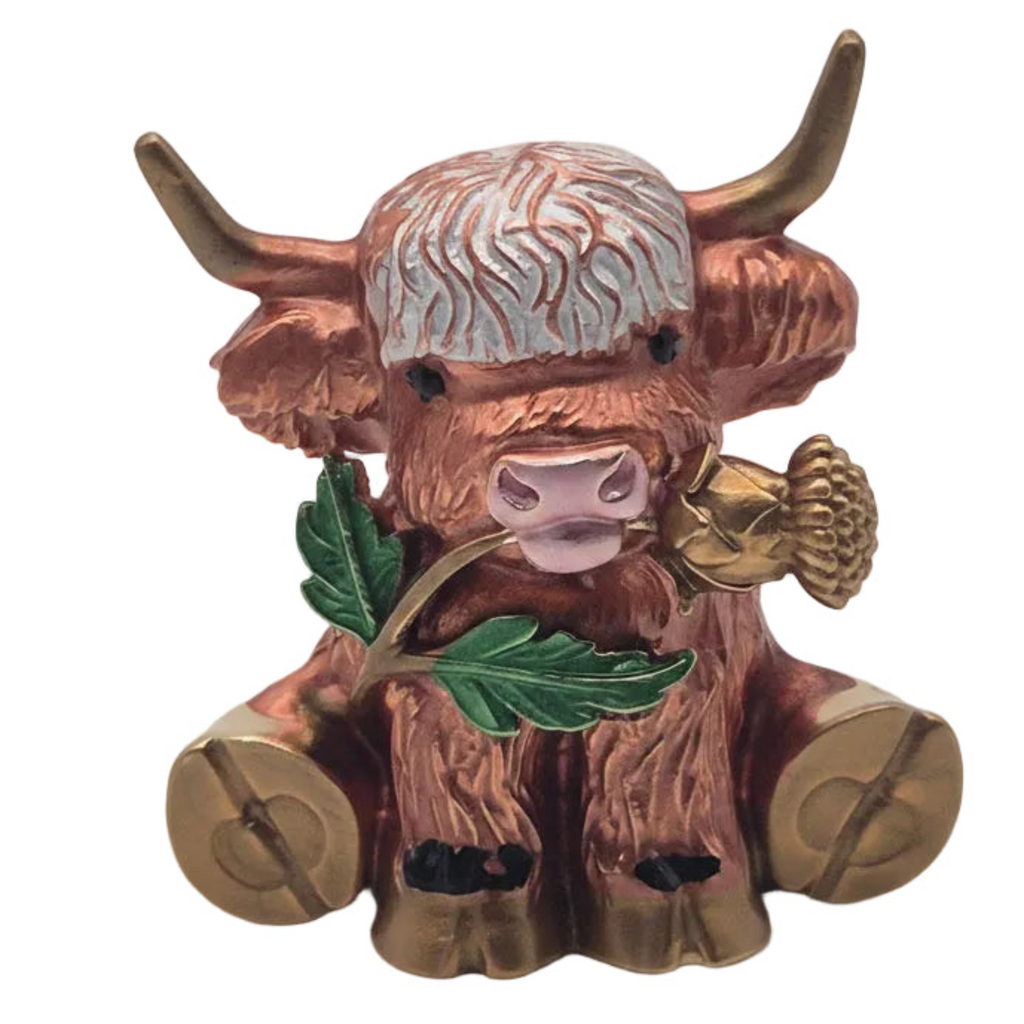A decorative figurine brooch of a Highland cow with copper-brown fur, gold-coloured horns and hooves, a white shaggy head, and green leaves in its mouth, set against a plain white background.