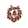 A gold-toned Christmas wreath brooch featuring red and black enamel leaves, a central gold bell, and clear rhinestones, shown on a plain white background.