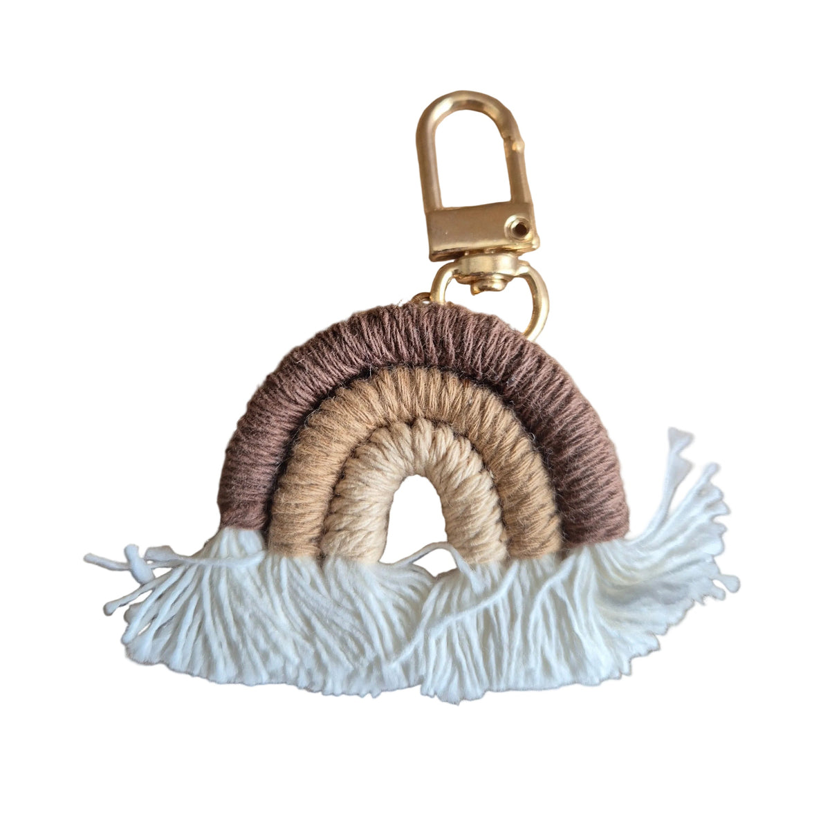 A handmade boho-style keychain featuring a small rainbow made of three arched layers of yarn in dark brown, tan, and beige, finished with a white fringed tassel and a gold metal clasp, displayed on a light brown wood grain surface.