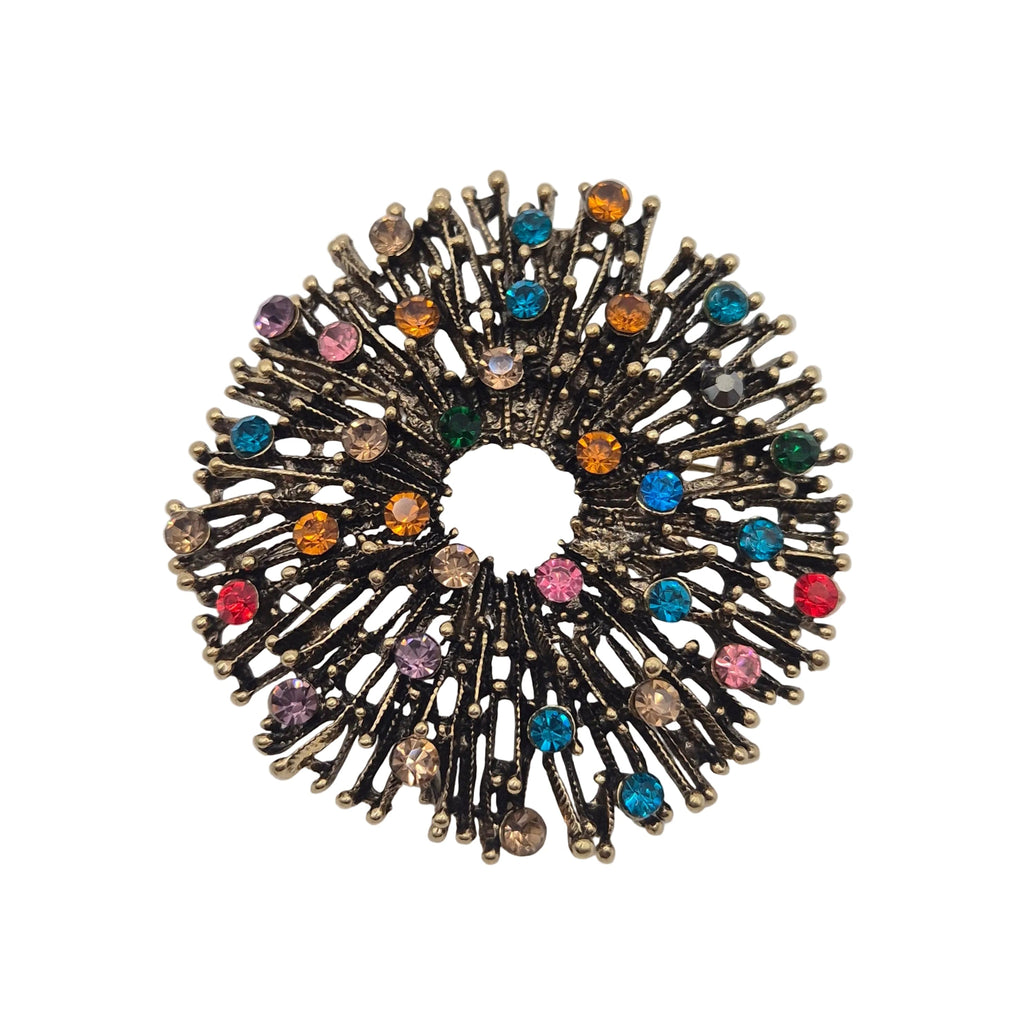 A circular firework brooch featuring a antique gold metal base with multiple radiating spikes adorned with small, colourful rhinestones in pink, blue, orange, green, and purple, set against a plain white background.