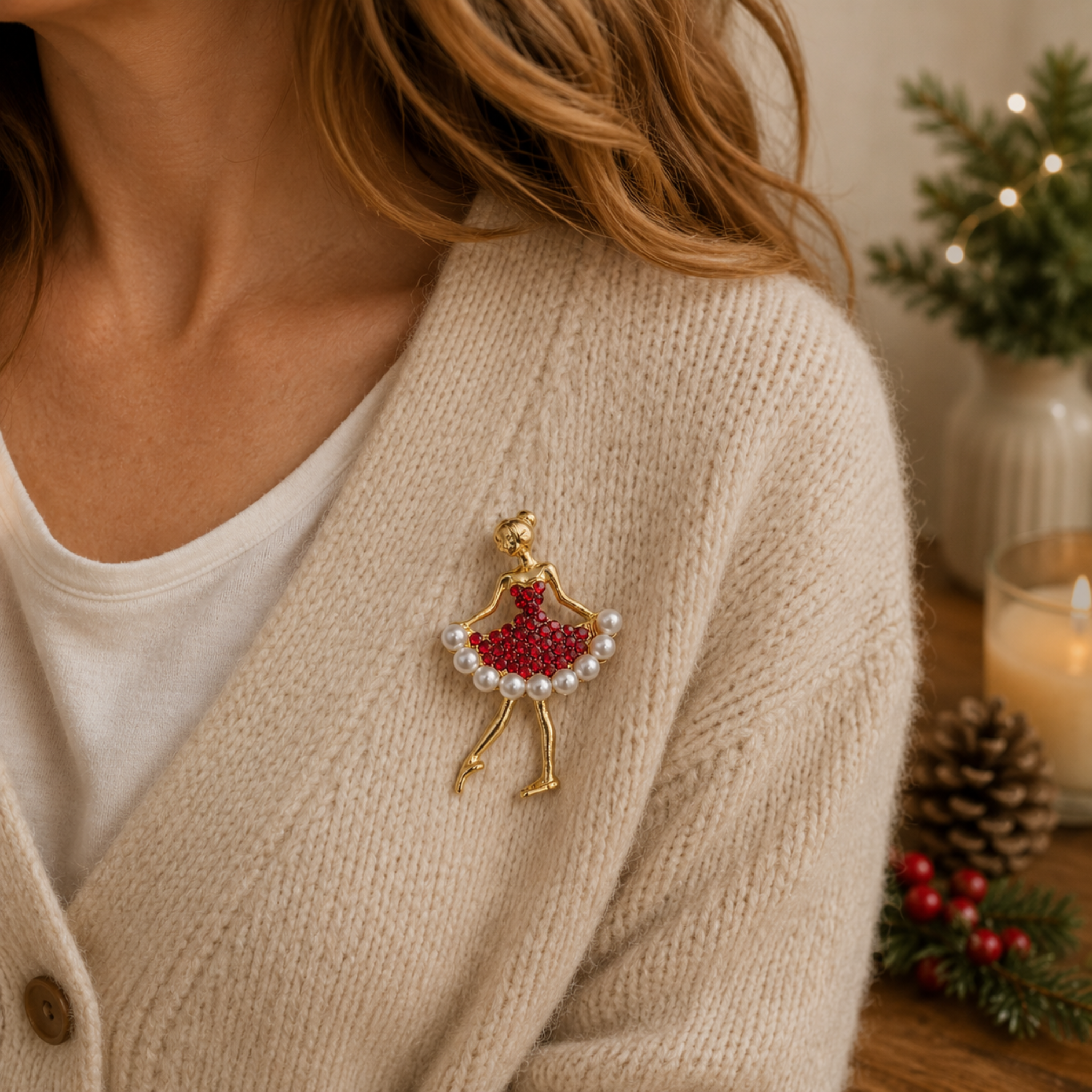A close-up lifestyle shot of a woman wearing a gold-toned brooch shaped like a dancing ballerina, adorned with a red beaded skirt and a border of white faux pearls. She is wearing a cream-colored knit cardigan over a white t-shirt, with a cozy holiday background featuring a lit candle, pinecone, and greenery.