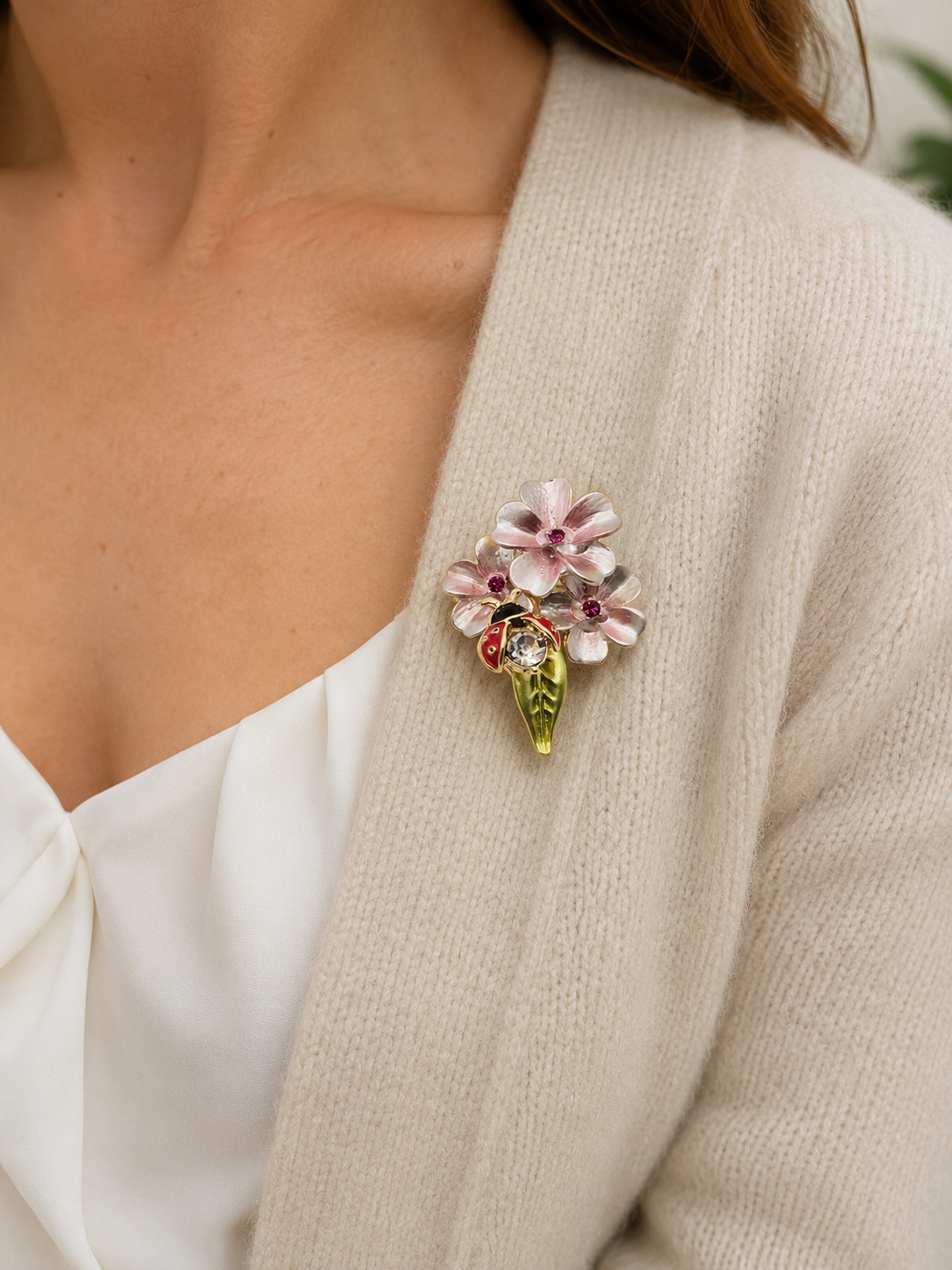 A close-up of a woman wearing a floral enamel brooch on her cream cardigan. The brooch features light pink and white flowers with green leaves and a small red accent.