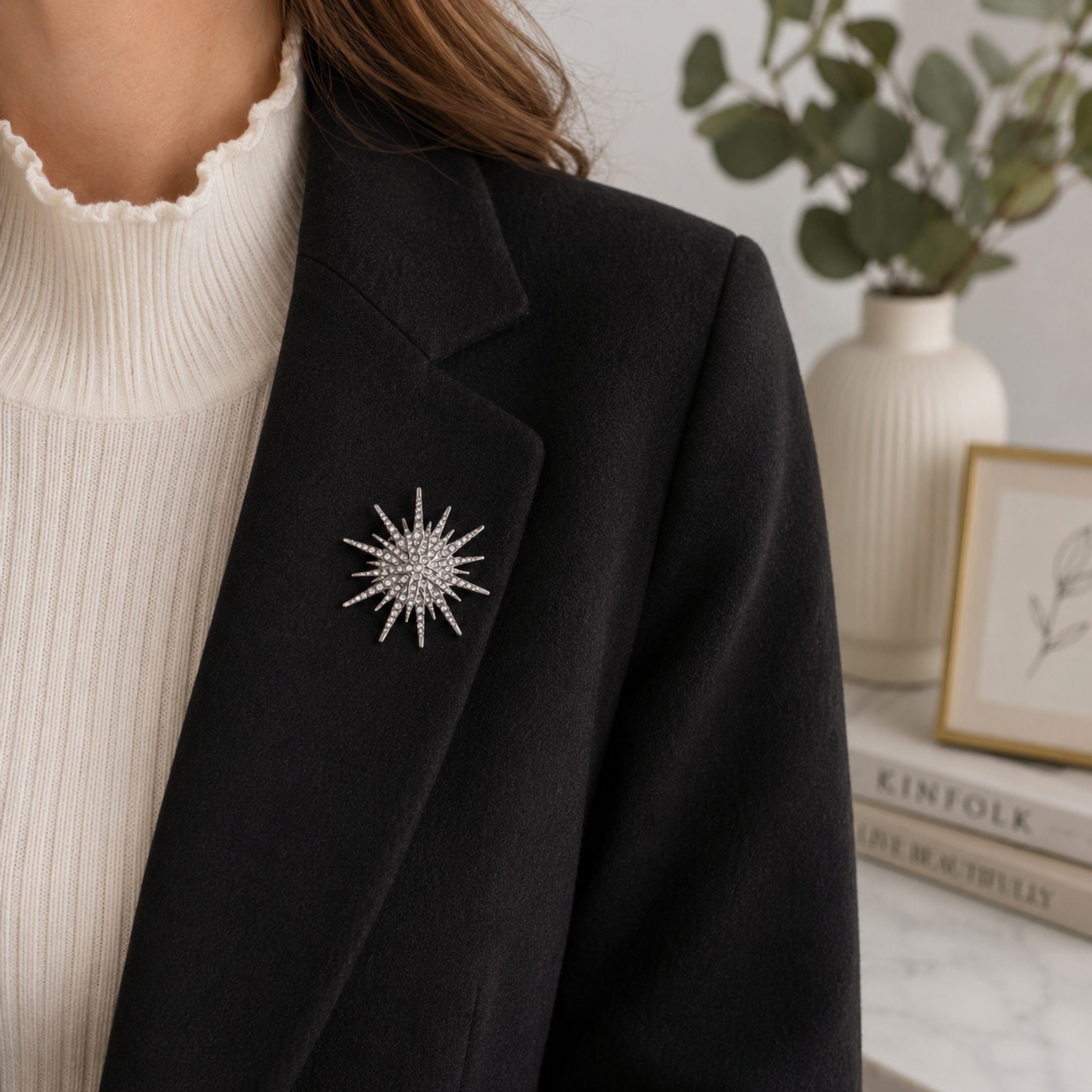 Silver toned metal starburst brooch pin, shown being worn on lapel of a black blazer. Vase of leaves and photo frame in the background.