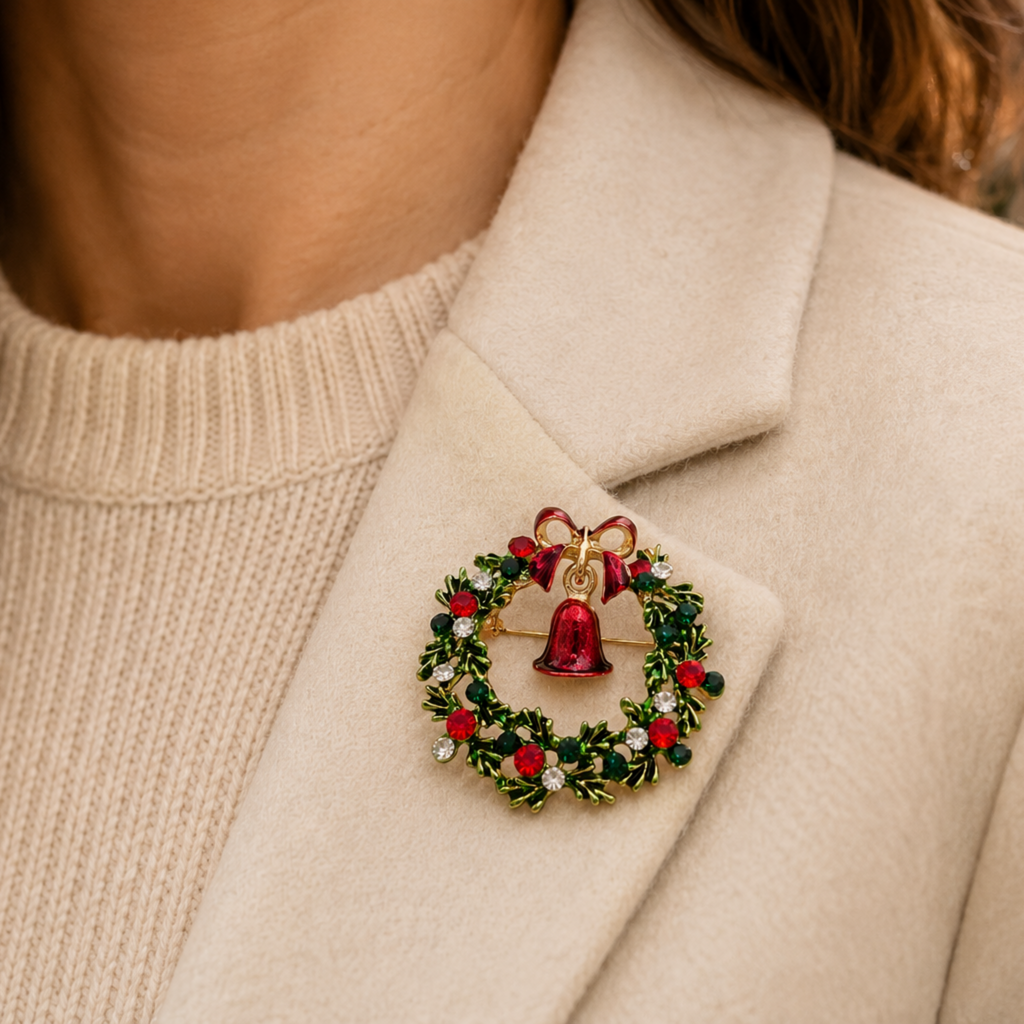 A close-up of a woman wearing a cream-colored coat and a ribbed cream sweater, featuring a festive Christmas wreath brooch pinned to the lapel. The brooch is detailed with green leaves, red berries, a red bow, and a gold bell.